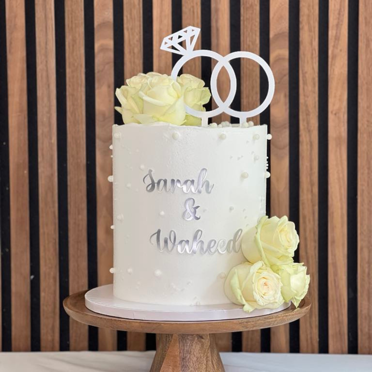 Timeless Romance tårta- Den sofistikerade Timeless Romance Wedding Cake är den perfekta bröllopstårtan för par som vill ha en klassisk och romantisk stil. Med vita rosor, pärldetaljer och eleganta ringdekorationer blir denna tårta en elegant mittpunkt på bröllopsdagen. Beställ din från Cocodrip för en tidlös och minnesvärd bröllopsfest! Rosorna är ej ätbara och plockas enkelt bort innan servering. Den oätbara caketoppern med ringarna ingår. ÄR DU ALLERGISK? KONTAKTA OSS OM ALLERGENER.040-104088 eller info@c